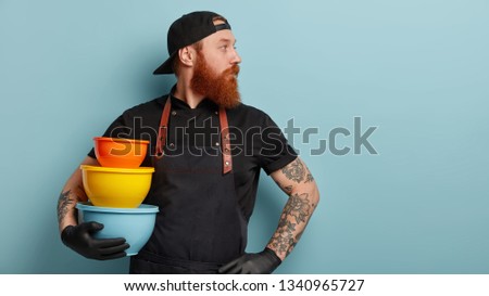 Indoor shot of serious man with thick foxy beard, focused aside with thoughtful expression, carries colorful food containers, poses indoor over blue background, works in restaurant at kitchen