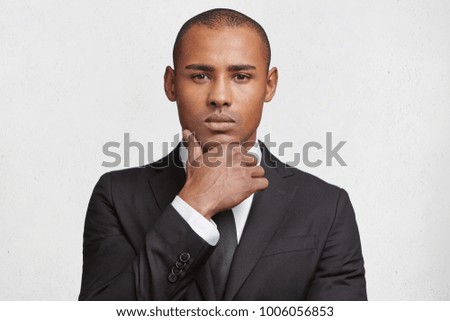 Indoor shot of serious mixed race male magnat dressed in elegant black suit, looks confidently into camera, waits for business partner as going to sign indemnity contract, isolated over white wall