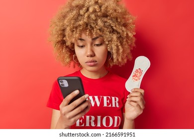 Indoor shot of serious woman checks menstruation calendar on smartphone holds sanitary napkin surfs information how to stop bleeding dressed in casual t shirt isolated over red vivid background