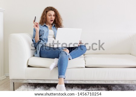 Indoor shot of shocked emotional lady with curly hair sitting in light living room, raising one hand, holding credit card, using her laptop, doing purchases on net, being fond of shopping online.