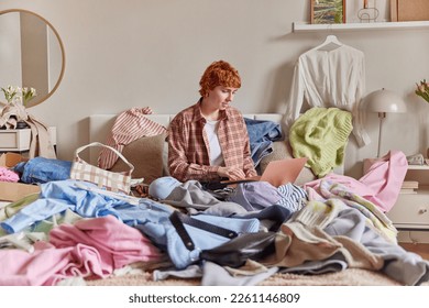 Indoor shot of short haired ginger woman surrounded by clothes from wardrobe sells different items of clothing online via laptop computer sits in bed against bedroom interior. Sorting and cleaning up