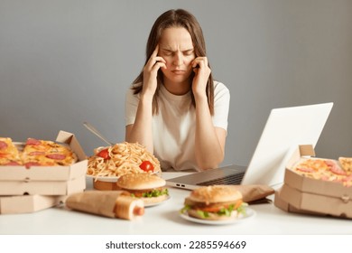 Indoor shot of sick unhealthy woman sitting in front of laptop among junk food, having terrible headache, being tire and exhausted, isolated over gray background.