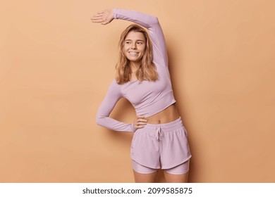 Indoor Shot Of Slim Active European Woman Has Workout Indoor Exercises In Gym Keeps Arm Raised Up Hand On Waist Leans Aside Dressed In Sportswear Smiles Happily Isolated Over Beige Background.