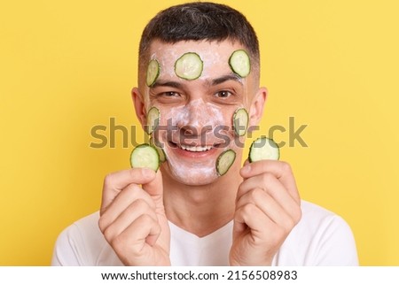 Indoor shot of smiling attractive man wearing white t shirt, posing with mask and slices of cucumbers on his face, expressing positive emotions, enjoying spa procedures.
