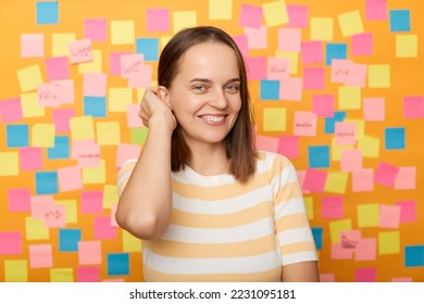 Indoor shot of smiling attractive woman with brown hair in striped t shirt standing against yellow wall with memo cards, looking at camera and smiling, touching her hair. - Shutterstock ID 2231095181