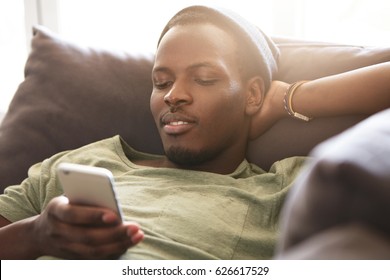 Indoor Shot Of Smiling Black Young Man Surfing Internet On Mobile Phone, Messaging Friends Online And Checking Newsfeed Via Social Media While Relaxing At Home, Lying On Couch With Hands Behind Head