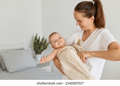 Indoor Shot Of Smiling Dark Haired Woman Wearing White T Shirt Standing With Her Baby Daughter In Hands, Kid Wrapped In Towel, Child Just After Taking Shower.