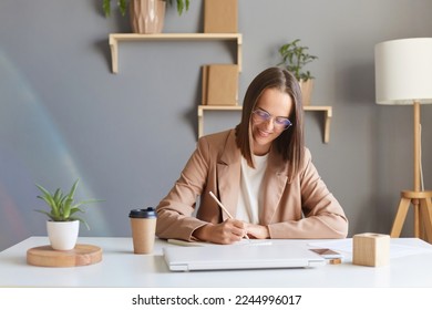 Indoor shot of smiling joyful woman freelancer or designer with brown hair wearing beige jacket working in office, drawing sketches or checking paper documents. - Shutterstock ID 2244996017