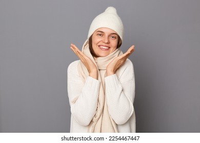 Indoor shot of smiling positive attractive woman wearing sweater, hat and scarf posing isolated over gray background, keeps palms near face, looking at camera with toothy smile, being in high spirit. - Shutterstock ID 2254467447