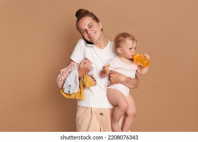 Indoor shot of smiling positive optimistic mother with baby daughter posing isolated over brown background, woman dressed in white t shirt, talking mobile phone, expressing happiness. - Powered by Shutterstock