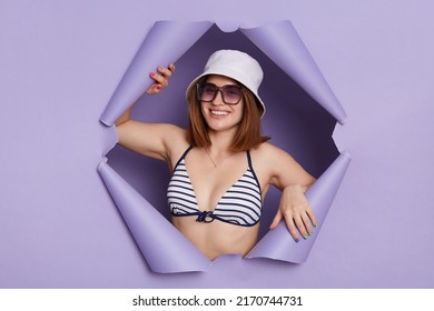 Indoor Shot Of Smiling Satisfied Woman Wearing Striped Swimming Suit Posing In Torn Purple Paper Wall, Looking At Camera With Positive Facial Expression, Being Happy.