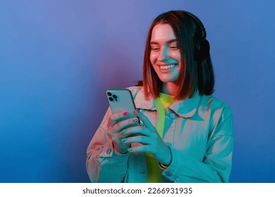 Indoor shot of smiling young girl wearing casual jacket and headphones standing isolated over blue neon light background, using her mobile [hone, chatting with friends, enjoying free time.