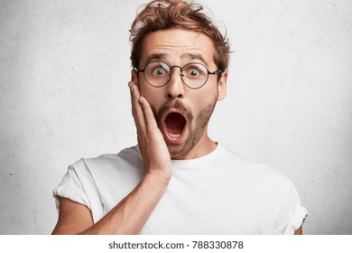 Indoor shot of stupefied bearded male professor wears round spectacles, keeps hand on cheek and keeps mouth widely opened, wonders about something, isolated over white concrete studio background
