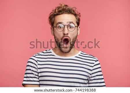 Indoor shot of stupefied handsome male opens mouth widely, wears striped t shirt, has shocked expression, being surprised, isolated over pink background. Astonished young crazy man feels stress