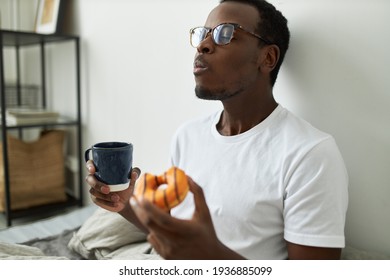 Indoor shot of stylish hungry young dark skinned man in glasses eating glazed orange doughnut with great relish, closing eyes with pleasure, enjoying every bite of sweet dessert, drinking morning tea