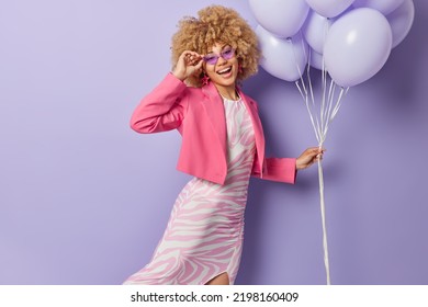 Indoor Shot Of Stylish Young Woman Wears Long Dress Pink Jacket And Sunglasses Holds Bunch Of Inflated Balloons Enjoys Party Time Isolated Over Purple Background. People And Holiday Concept.