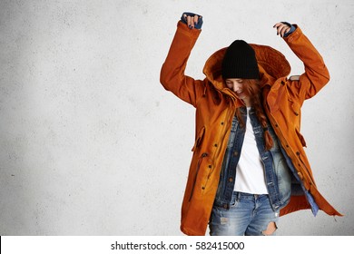 Indoor shot of stylish young woman model wearing fashionable red winter coat, black hat and ragged jeans having fun, lifting her hands up while posing isolated against grey studio wall background