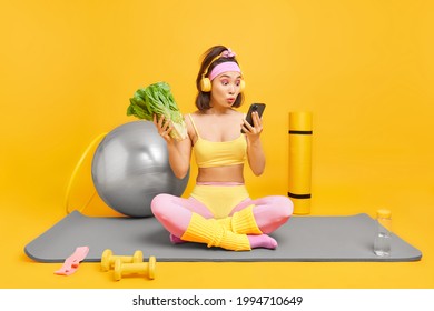 Indoor shot of surprised Asian woman looks shocked at smartphone display sits crossed legs on fitness mat chooses song from playlist leads active lifestyle keeps to healthy diet poses at home gym