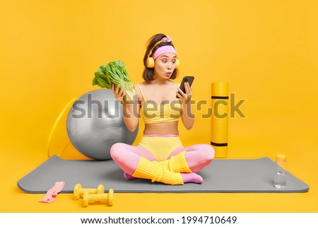 Indoor shot of surprised Asian woman looks shocked at smartphone display sits crossed legs on fitness mat chooses song from playlist leads active lifestyle keeps to healthy diet poses at home gym