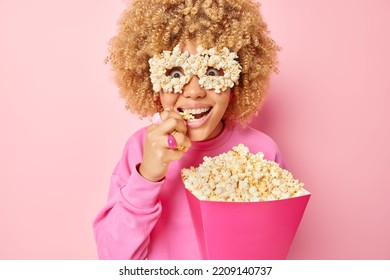 Indoor shot of surprised curly haired woman grabs delicious popcorn from paper bucket watches very intersting mobie excited by breathtaking scene dressed casually isolated over pink background