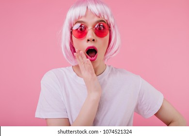 Indoor shot of surprised female model in short periwig and bright sunglasses. Portrait of shocked young woman in peruke isolated on pink background.