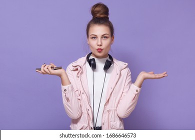 Indoor Shot Of Teenager Caucasian Woman Wears White Shirt And Pink Jacket, Posing Isolated Over Lilac Wall, Looks Directly Atcamera, Makes Kiss Gesture, Spreads Palms Aside, Has Headphone Around Neck.