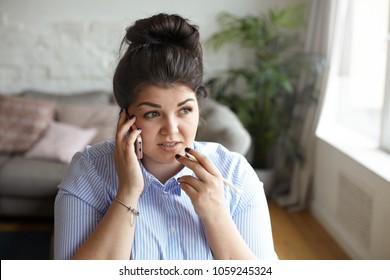 Indoor shot of thoughtful plus size young brunette businesswoman dressed in blue shirt making business call at home office, talking to her accountant while doing finances, having pensive look - Powered by Shutterstock