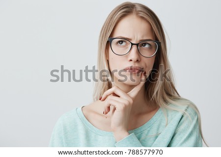 Indoor shot of thoughtful pretty woman has long blonde hair with stylish eyewear, looks aside with pensive expression, plans something on coming weekends, poses against blank wall. Puzzled female