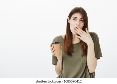 Indoor Shot Of Tired And Bored Cute Girlfriend In Dark-green T-shirt, Yawning, Covering Opened Mouth With Palm, Drinking Coffee Not To Fall Asleep During Daytime, Overworking Or Hearing Boring Talks