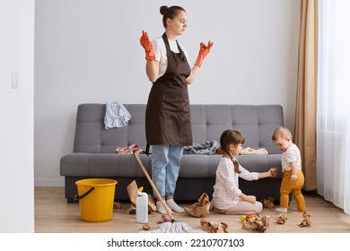 Indoor Shot Of Tired Female With Bun Hairstyle Washing Floor With Mop At Home, Children Making Mess In Living Room, Exhausted Mother Trying Calm Down And Relax.