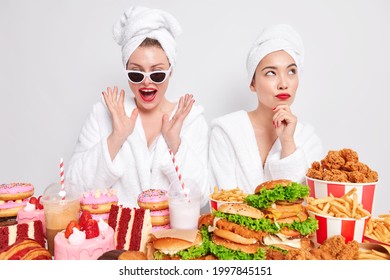 Indoor shot of two women have cheat day spend free time at home stand near delicious snacks wear bath towel and robe eat tasty burgers cakes donuts drink fizzy drinks. Delicious unhealthy food