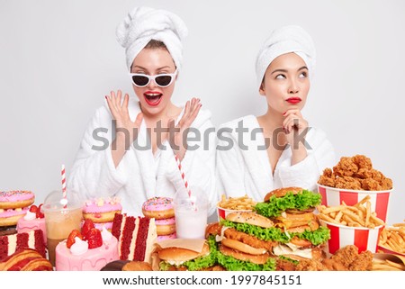 Indoor shot of two women have cheat day spend free time at home stand near delicious snacks wear bath towel and robe eat tasty burgers cakes donuts drink fizzy drinks. Delicious unhealthy food