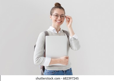 Indoor Shot Of University Student Girl Standing Isolated On Gray Background With Closed Laptop In Hand, Setting Eyeglasses Straight With Open Positive Smile, Expressing Enthusiasm And Initiative.