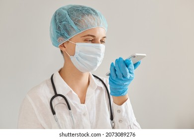 Indoor Shot Of Woman Doctor Wearing Medical Cap, Gloves And Protective Mask Using Mobile Phone, Recording Voice Message, Expressing Positive Emotions And Happiness.