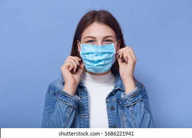 Indoor shot of woman wearing disposable protective face mask. Air pollution, industrial emissions or virus epidemic concept, female wearing denim jacket and white shirt stands over lilac studio wall. - Powered by Shutterstock