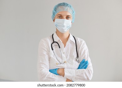 Indoor Shot Of Young Adult Caucasian Doctor Woman Wearing Gloves, Medical Mask And Cap, Gown, Standing With Folded Hands And Looking At Camera, Working In Hospital Or Clinic.