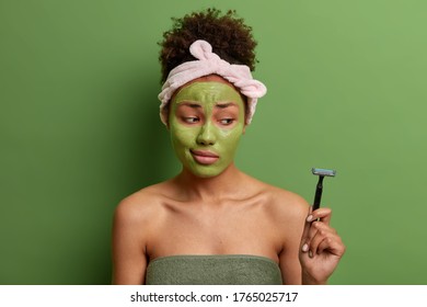 Indoor Shot Of Young Adult Woman With Afro Hair Applies Green Beauty Mask Against Facial Wrinkles, Uses Cosmetic Product For Beauty, Rejuvenation And Skin Care Holds Razor Isolated On Green Background