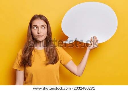 Indoor shot of young beautiful european disappointed female with long loose straight hair wearing casual tshirt isolated on yellow background holding white speech bubble for your advertisement