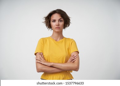 Indoor shot of young brunette lady with short haircut keeping her hands folded on chest, looking at camera and raising eyebrow confusedly, isolated over white background