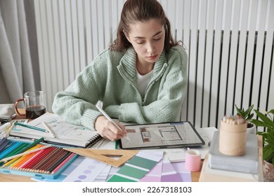 Indoor shot of young female interior designer works on apartment design project draws in digital tablet with stylus thinks about decorations of apartment wears casual jumper poses at desktop. - Powered by Shutterstock