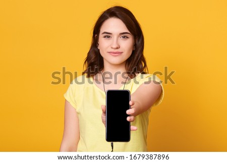 Indoor shot of young happy attractive european female with smartphone in hands, showing its bolank screen, posing isolated over yellow background, listening to music via earphones. Copy space.