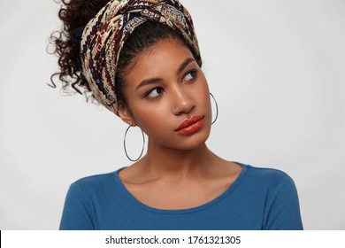 Indoor shot of young pretty dark haired curly lady tilting her head while looking wonderinly aside, dressed in blue blouse and colored headband while posing over white background