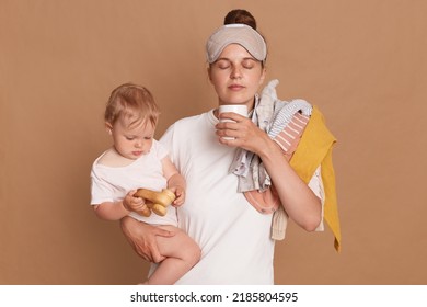 Indoor Shot Of Young Tired Mom And Little Child After Sleepless Night, Exhausted Woman With Baby Standing With Coffee Isolated Over Brown Background. Postpartum Depression On Maternity Leave.