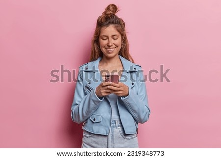 Indoor shot of young woman explores social media content on her smartphone seamlessly connected to internet eagerly downloading new application dressed in blue jacket isolated on pink studio wall