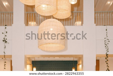 An indoor space with a high ceiling, decorated with several large woven pendant lights. Multiple dome-shaped, basket-woven rattan chandeliers hang at varying heights. Large white rectangular columns and a second-story balcony with white railings create a structured backdrop.