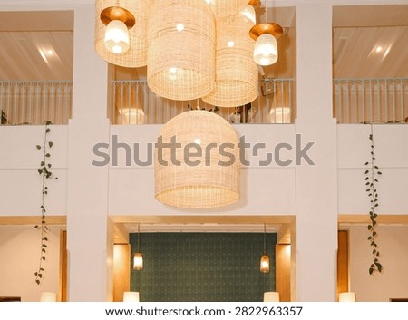 An indoor space with a high ceiling, decorated with several large woven pendant lights. Multiple dome-shaped, basket-woven rattan chandeliers hang at varying heights. Large white rectangular columns and a second-story balcony with white railings create a structured backdrop.