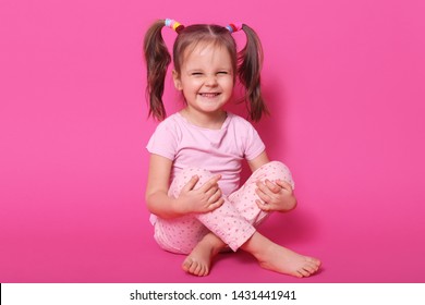 Indoor Studio Shot Of Laughing Positive Kid Sitting On Floor, Posing Isolated Over Pink Background, Wearing Rose T Shirt And Trousers, Having Ponytails, Being In High Spirits. Childhood Concept.