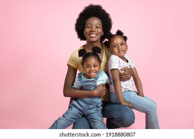 Indoor studio shot of pleasant young African woman mother and her two cute adorable little daughters, wearing bright trendy casual clothes, posing together on pink background. Motherhood