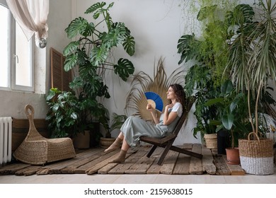 Indoor Tropical Oasis. Young Relaxed Dreamy Woman Using Paper Fan While Escaping Summer Heat In Beautiful And Refreshing Home Garden Full Of Large-leaved Exotic Plants, Dreaming About Vacation