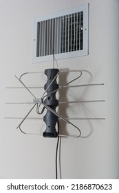 Indoor TV Antenna Hanging By Wire Coat Hanger From Heating Duct On White Textured Wall. Oblique Angle View From Wall